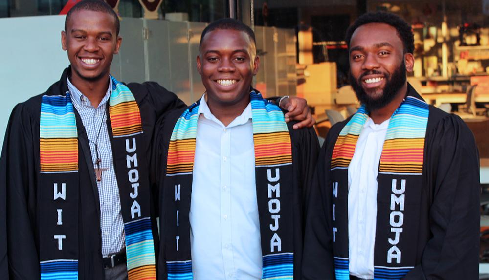 Three black students in graduation robes wearing UMOJA scarves.