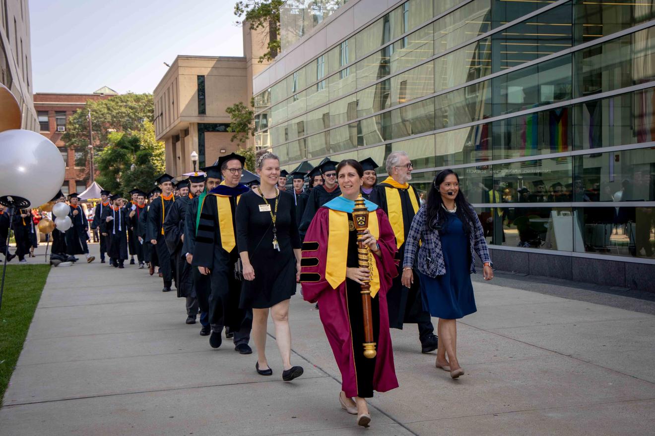Woman in a maroon robe carrying a brown pole leading men and women wearing black caps and gowns at their graduation ceremony.