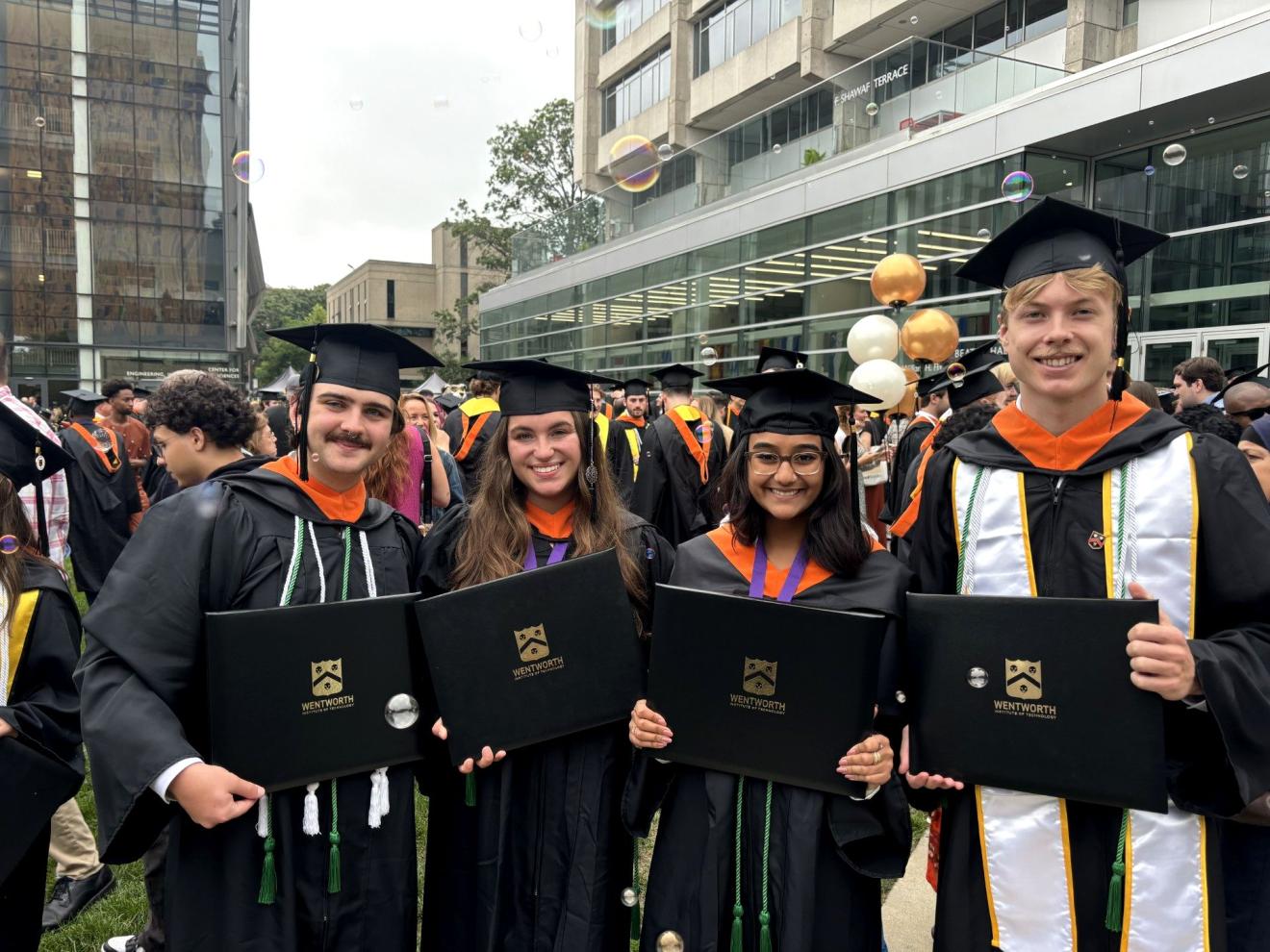 Group of smiling women & men wearing black caps and gown with orange hoods holding diploma covers after their graduation ceremony