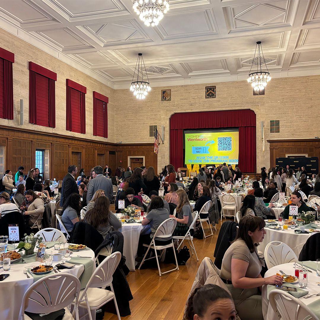 View of hall filled with tables and chairs and people attending a breakfast event looking at the screen and speaker at a podium