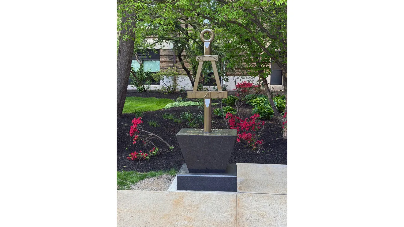a metal statue sits in a quad surrounded by trees and flowers