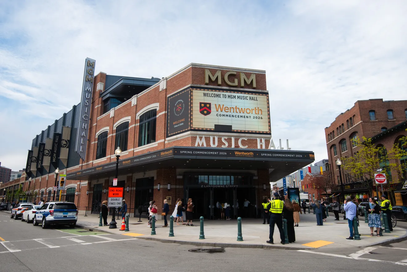 Marquee at MGM Music Hall where commencement is now held - brick building - corner view with cars lining the left including a Boston Police Car and view of the right side oof the street with families waiting to go in - several trees in bloom.