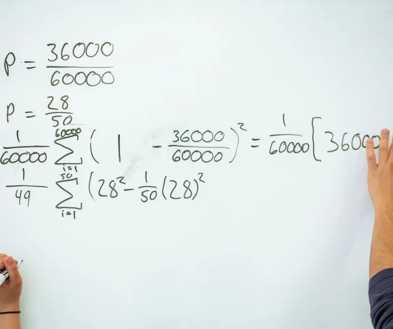 Two hands work on a math problem on a white board