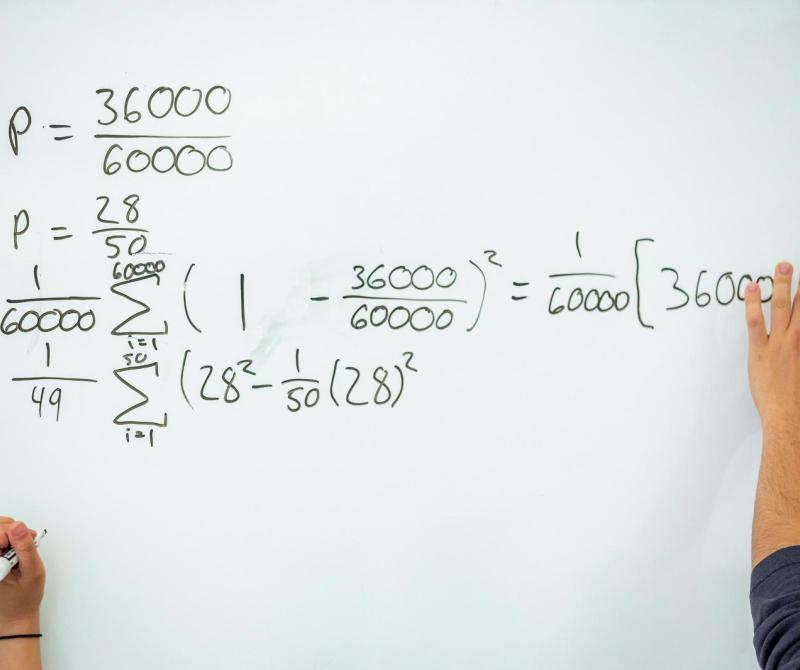 Two hands work on a math problem on a white board