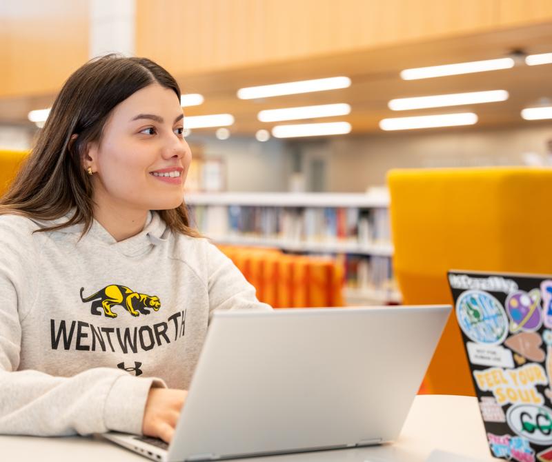 A student smiles over while working on her laptop in the Wentworth library