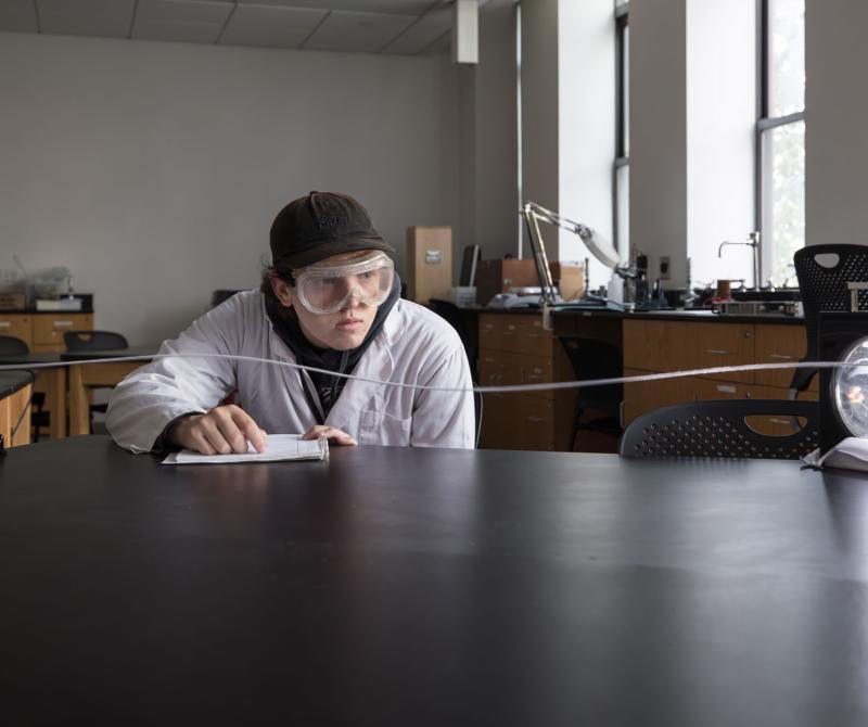 A male student wearing a lab coat and protective goggles conducts a period and frequency experiment in the Optics Lab