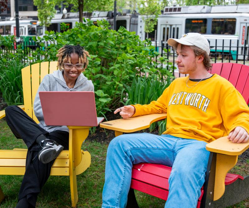 A Black female student and a white male wearing a Wentworth shirt sit together on the quad to study looking at a laptop. The Green Line T is in the background.