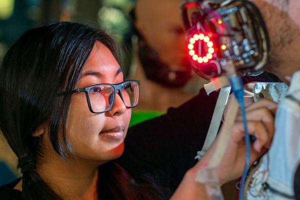 A woman in glasses adjust the glowing mask on the man modeling her costume.