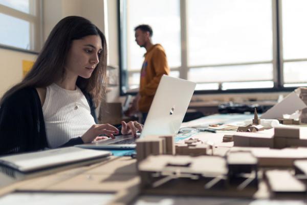 A female student works on a laptop surrounded by paper models in the architecture department. 