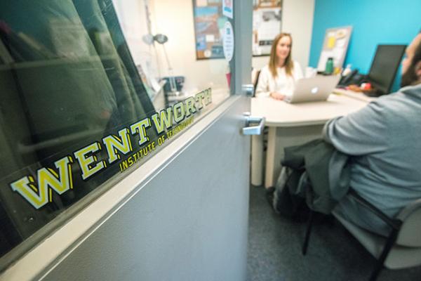 In the foreground, the focus is on a Wentworth decal on an office door. In the background a student sits across from a female advisor at her desk.