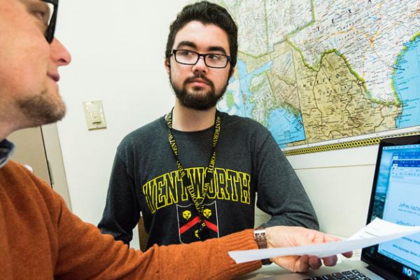 A male student looks on as an advisor talks to him in front of his laptop.
