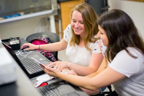Two female students sit in front of computers looking at an handheld device together. 