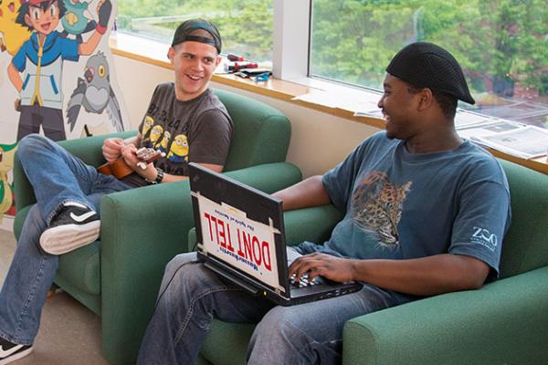 Two male students sitting next to each other and chatting. The student on the left plays the ukulele while the student on the right works on his laptop