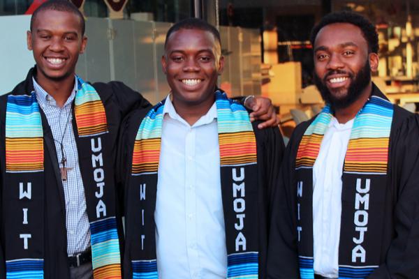 Three black students in graduation robes wearing UMOJA scarves.