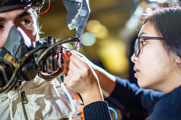A young woman adjusts the mask she designed on a male model.