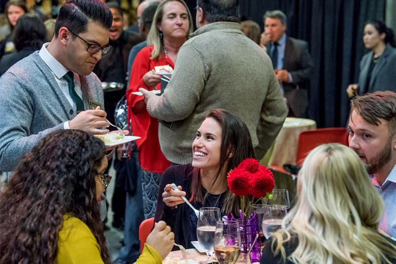 A group of faculty and staff, seated and standing, talk and eat at an event.