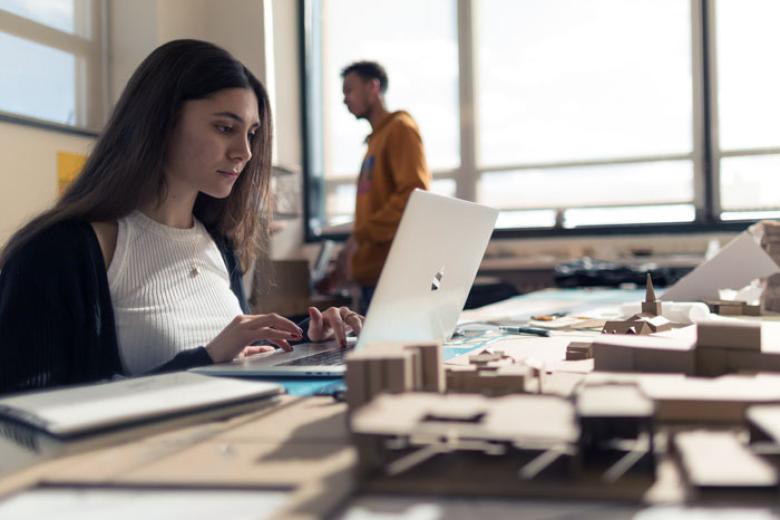 A female student works on a laptop surrounded by paper models in the architecture department. 