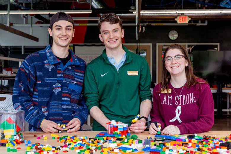 Two male students and one female student smile at the camera in front of a table of legos. 
