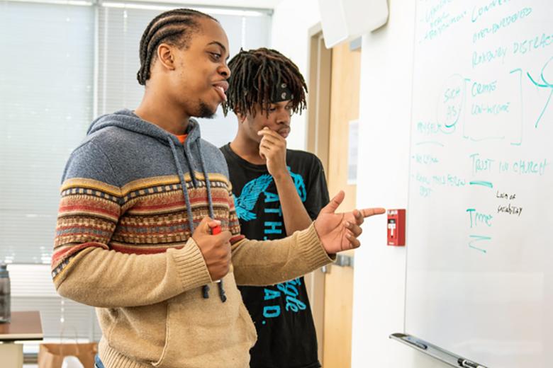 Two male, black students working at a whiteboard. 