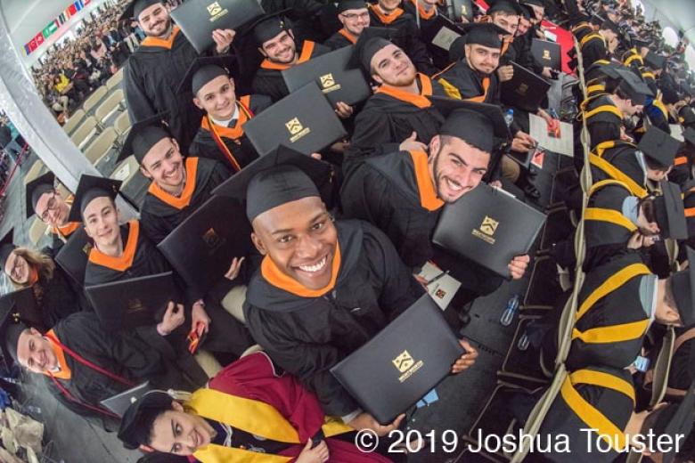 Group of students, white and brown, wearing their graduation cap and gown, smiling up and showing their diploma covers