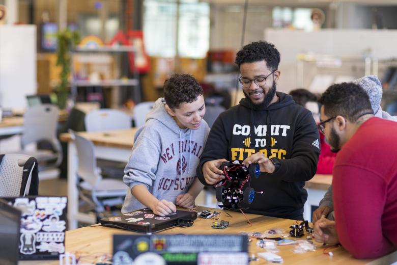 Three students brainstorm together around a table while holding a small robotic car