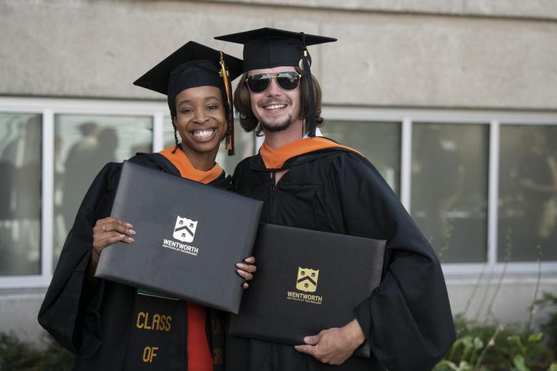 A female and male student pose and smile with their graduation caps and gowns and diplomas