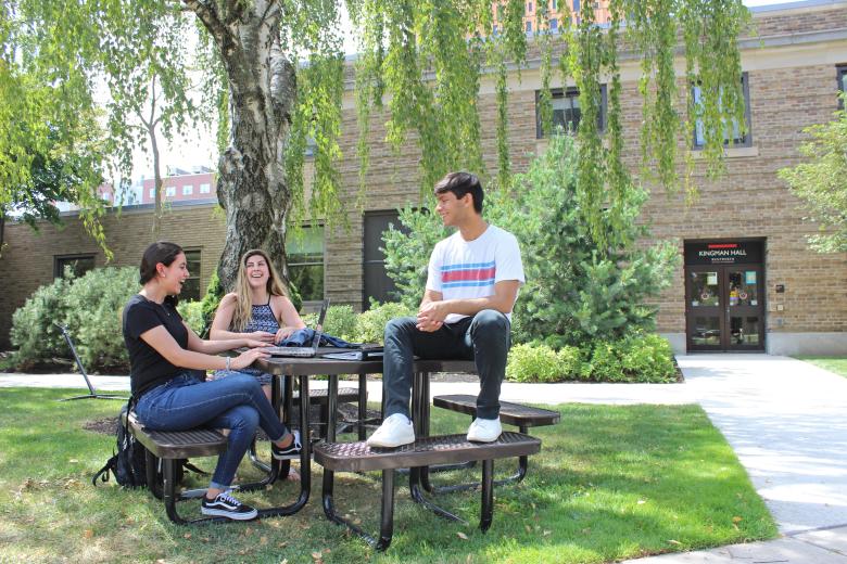 Three students sit at a picnic table in the quad while talking and smiling at one another