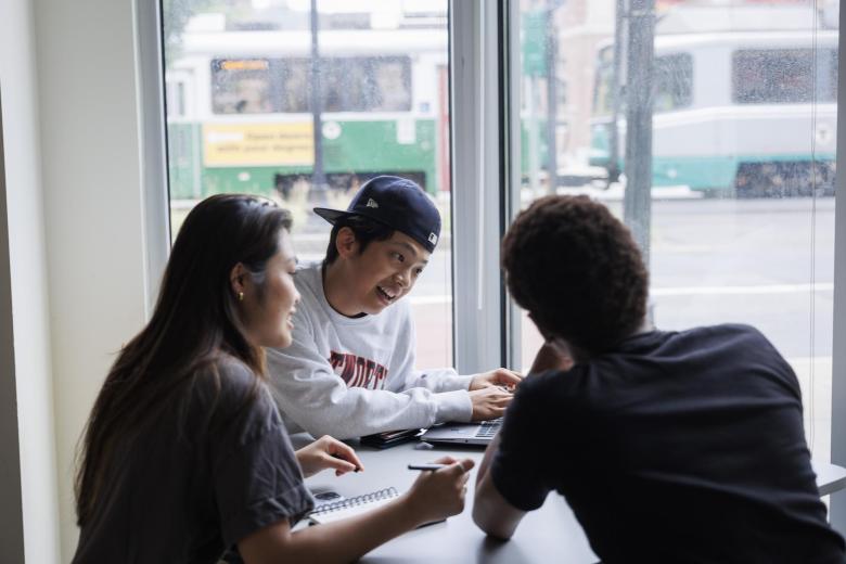 Three students discuss topics while sitting around their laptops