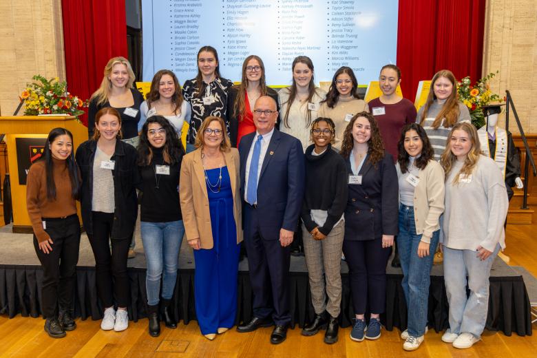 Group photo of female identifying students in 2 rows - all ages, heights, and one man (President Thompson) wearing a suit and glasses