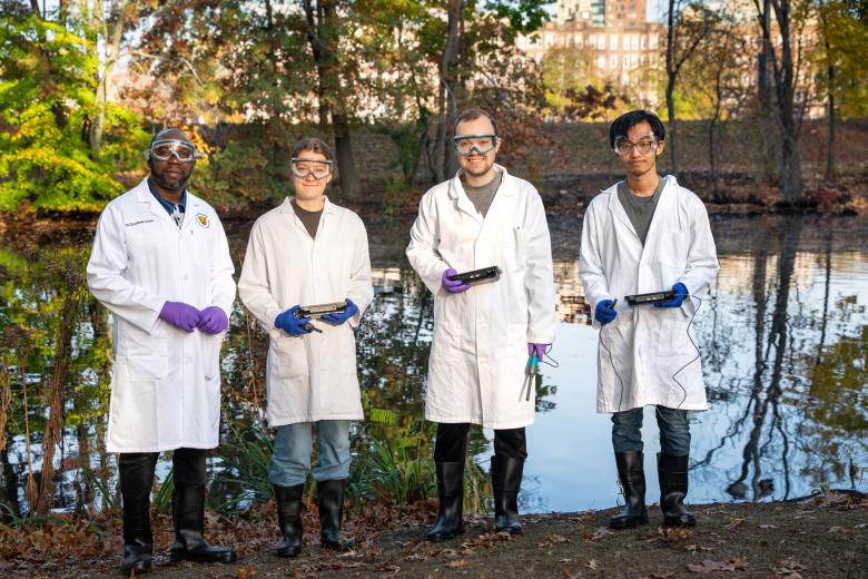 A faculty member and three students wearing lab coats stand by a body of water for measurements.