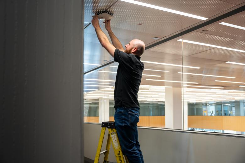 A person standing on a step ladder adjusting a Wi-Fi device on the ceiling.