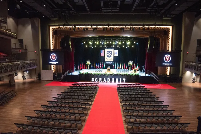 View of stage for commencement - red carpet flanked by black chairs for student seating, white cut out letters #graduwit, yellow, red and green florals decorating the black stage front, 26 international flags on poles representing the countries of international students, large Wentworth banner in gold and black with our school seal flanked by the US and Massachusetts state flags, 2 podiums and 35 black padded chairs on the stage for faculty and trustees
