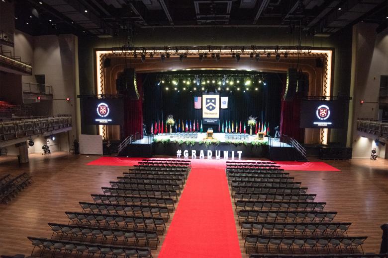View of stage for commencement - red carpet flanked by black chairs for student seating, white cut out letters #graduwit, yellow, red and green florals decorating the black stage front, 26 international flags on poles representing the countries of international students, large Wentworth banner in gold and black with our school seal flanked by the US and Massachusetts state flags, 2 podiums and 35 black padded chairs on the stage for faculty and trustees