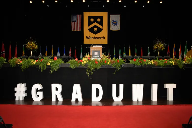 Stage with 26 flags representing countries of graduating students, 2 brown podiums with Wentworth logo, red and gold flowers along stage edge with greenery, large US Flag and Massachusetts State flag flanking the main Wentworth Gold and Black banner. Cut out letters #Graduwit in front of the black stage panel in front of a red carpet.