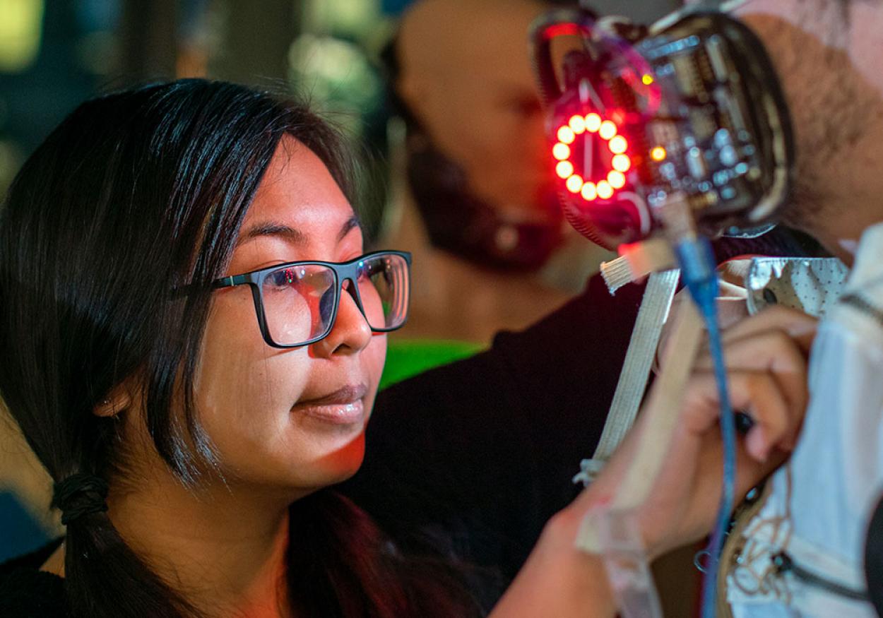 A woman in glasses adjust the glowing mask on the man modeling her costume.