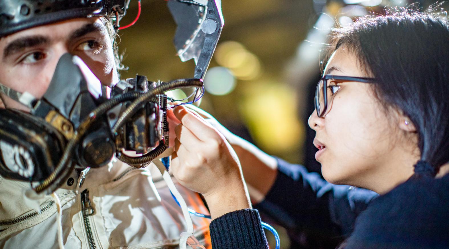 A young woman adjusts the mask she designed on a male model. 