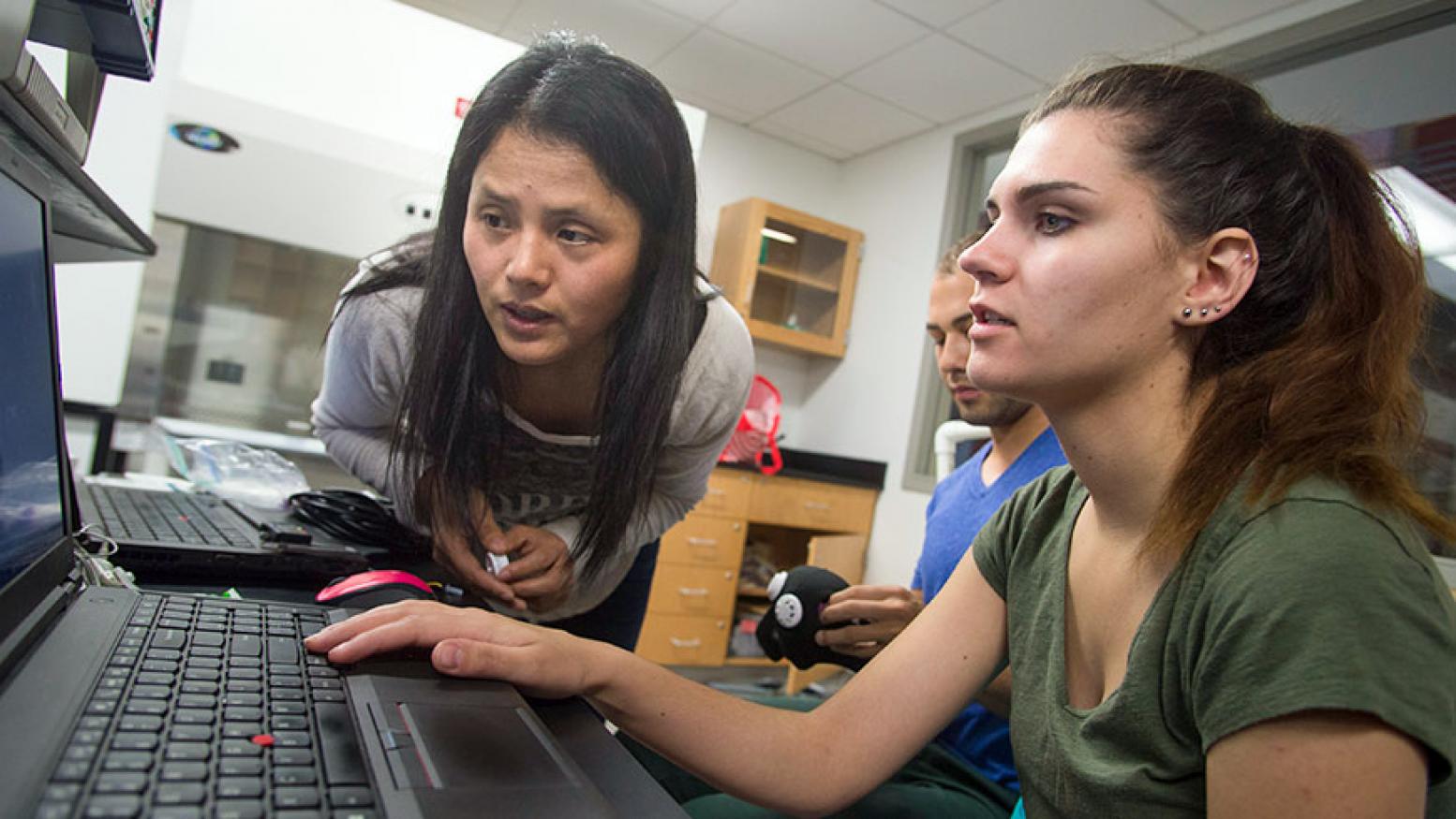 A female students looks at a computer while the professor leans over and examines her work. 