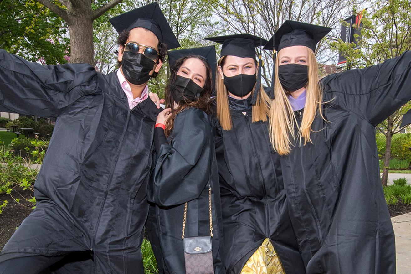 A group of 4 graduates, in caps and gowns wearing face masks, celebrate the day of commencement