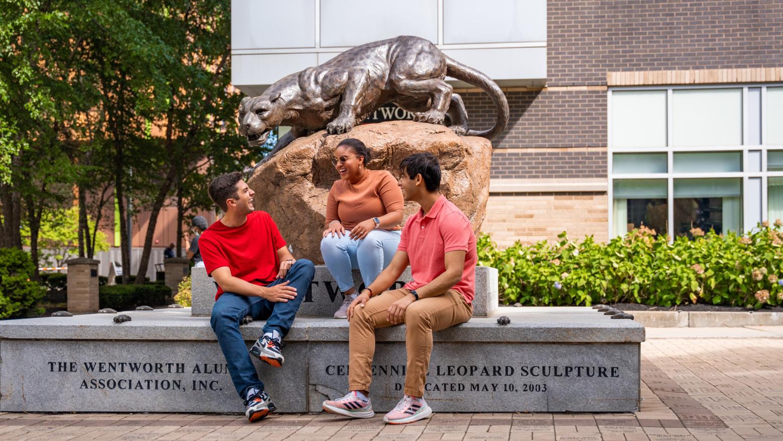 Three students smile and laugh in front of the Wentworth campus statue of a leopard