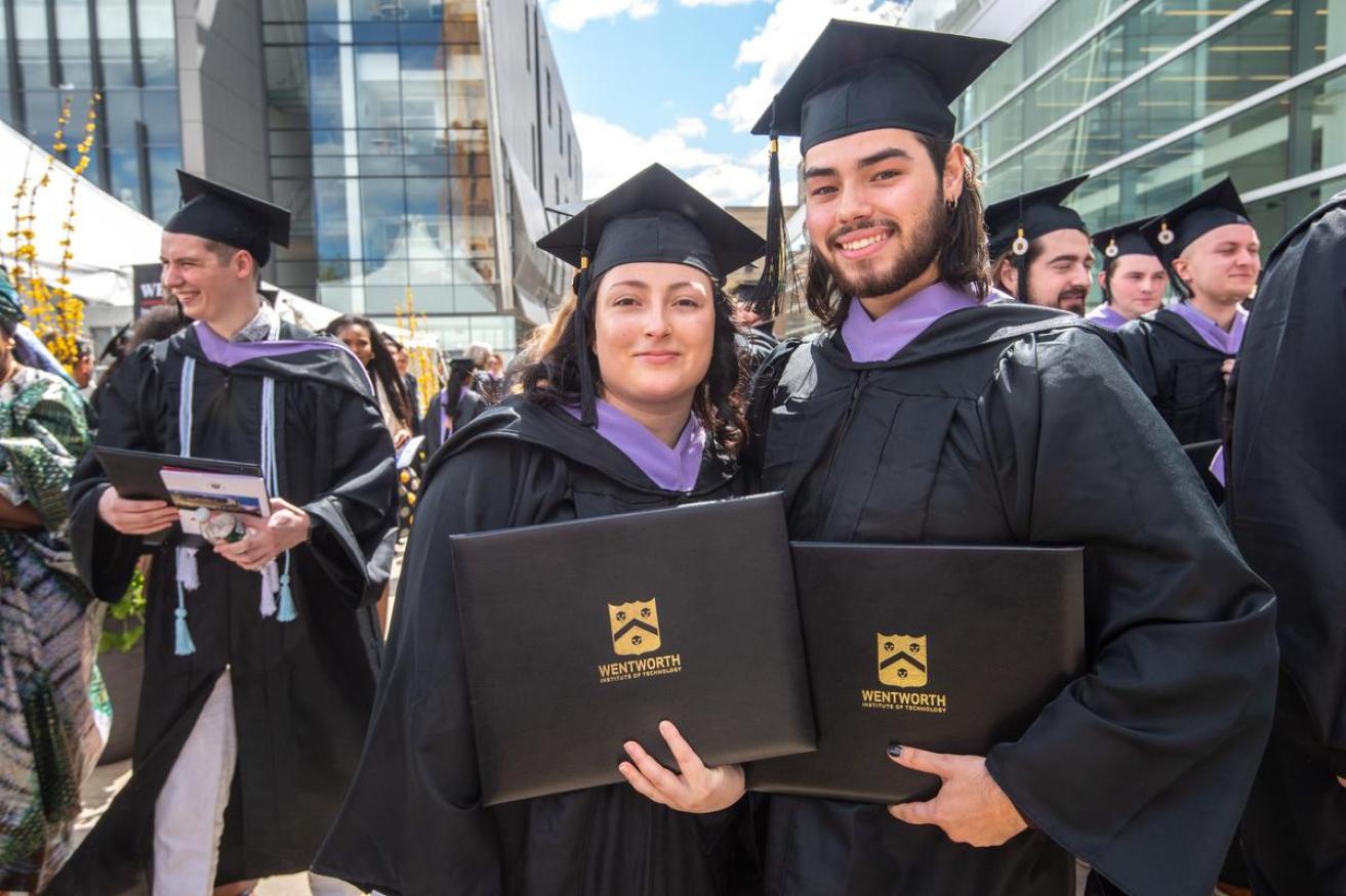 A male and female student pose for the camera at graduation holding up their diplomas and smiling