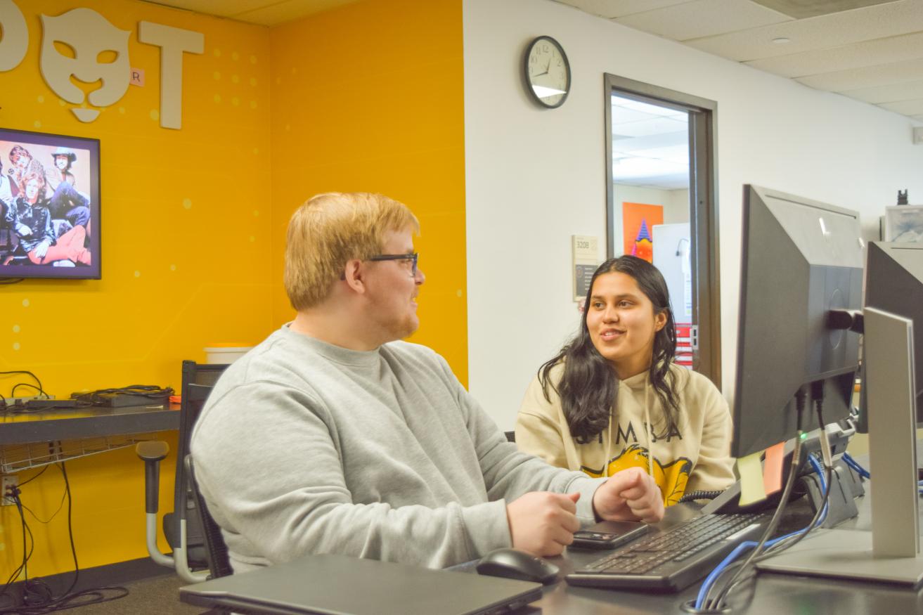 Two people behind a desk talking and Infront of a computer