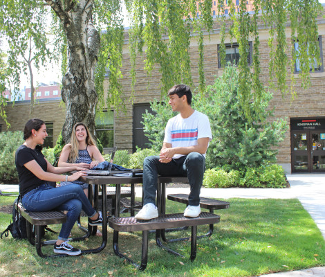 Three students sit at a picnic table in the quad while talking and smiling at one another