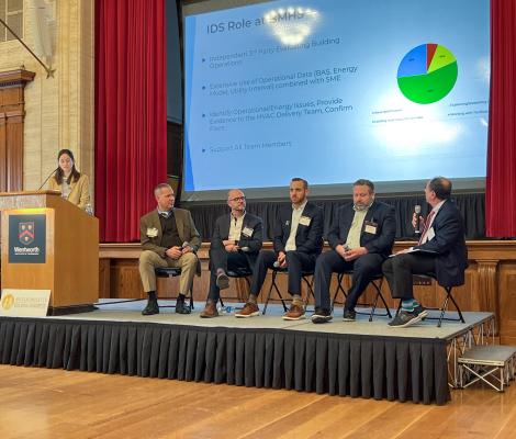 four people sit on a stage as part of a panel while a moderator stands at a podium