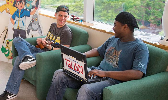 Two male students sitting next to each other and chatting. The student on the left plays the ukulele while the student on the right works on his laptop
