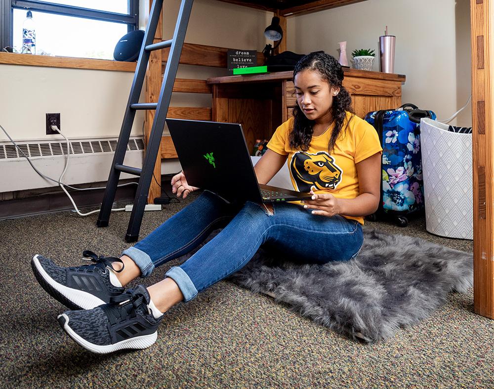 Students sits on floor of residence hall room and works on laptop.