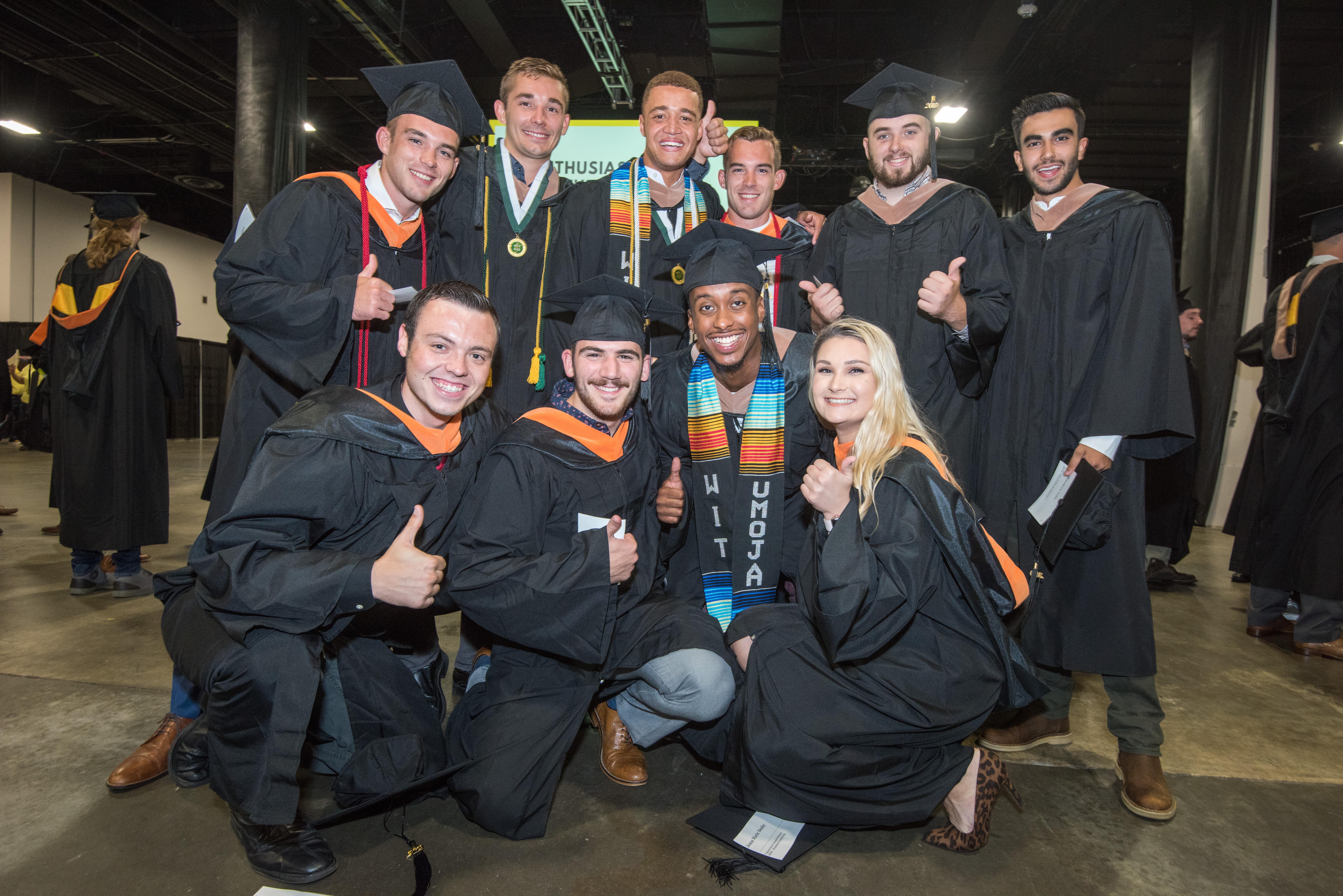 Group of men and a woman posing for a photo at commencement wearing their caps and gowns showing thumbs up.