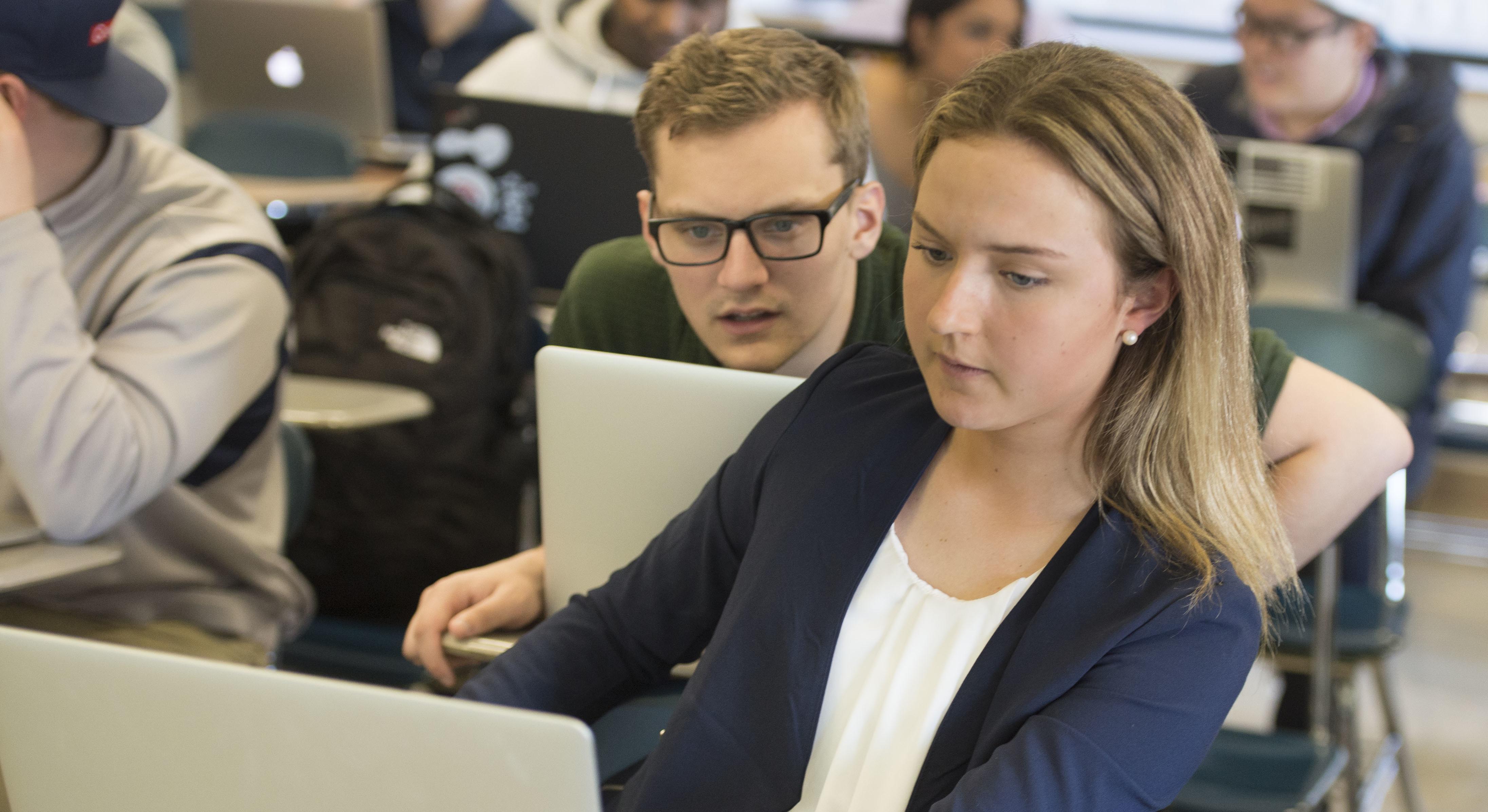 Image of two students in classroom. One is looking over the shoulder of the other at a laptop computer.