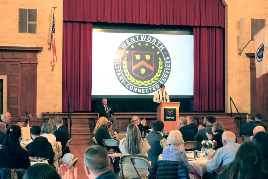 Student speaks out from a podium to several round tables filled with guests. 