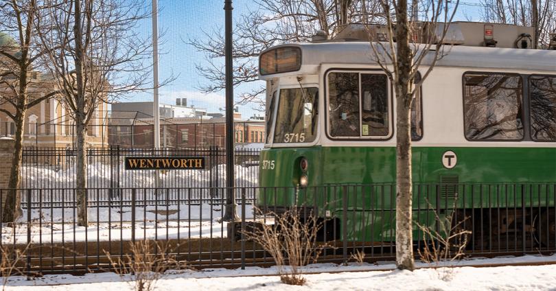 A Green Line train car passes by Wentworth Institute of Technology with the Wentworth sign prominent in the foreground