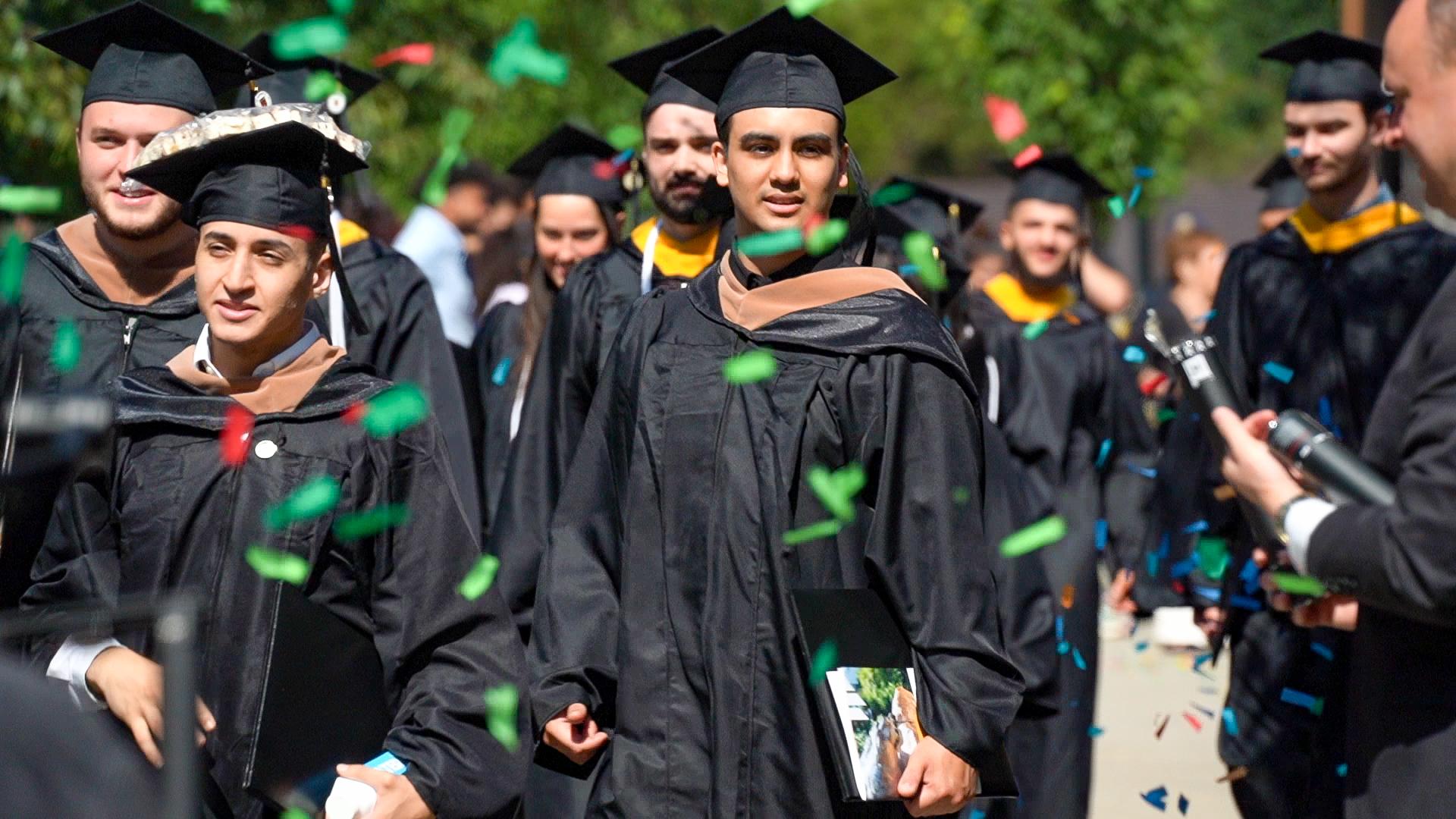 students wearing graduation robes walk through falling confetti